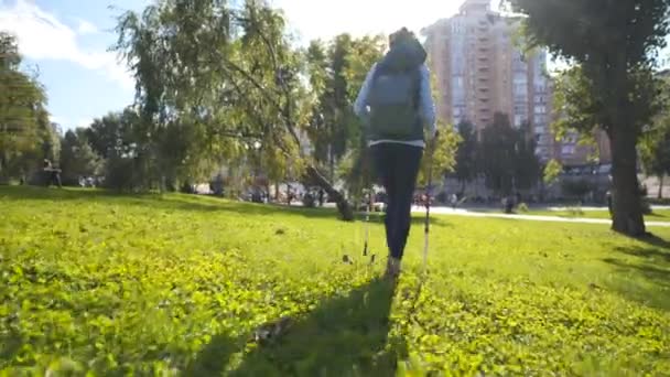 Egészséges és aktív. Felnőtt nő sétapóznákkal sétál ki a gyepre a városi parkban napsütésben. Vidám idős hölgy a zöld parkban, aki skandináv sétákat tesz. Skandináv pálcikák — Stock videók