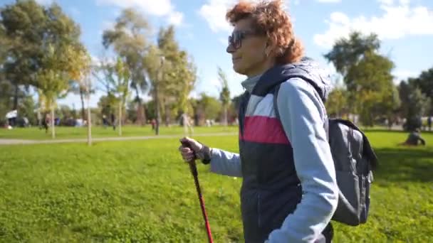 Femme mûre marchant dans le parc avec des bâtons de marche nordique par temps ensoleillé. Senior entraînement féminin scandinave marche dans la nature. Ajustement aux personnes âgées sportive exercice cardio avec des bâtons dans les mains en plein air — Video