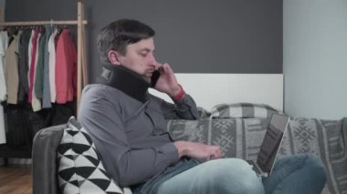 Boyunluk takan adam tele-telden çalışıyor. Köpüklü boyun tasması takmış yaralı bir erkek telefonda dizüstü bilgisayarla konuşuyor kanepede oturuyor ve boynu ağrıyor.