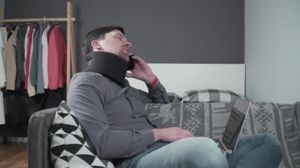 Man freelancer in cervical collar neck brace working from home. Male employee after accident working at laptop, talking on phone at couch. suffering from neck pain. Guy with spinal injury is calling — Stock Video