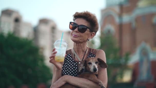 Vacances d'été de femme âgée solitaire et son ami chien parmi les passants foule gens sur la promenade. Boire une boisson froide avec de la glace de paille et une tasse transparente avec un animal de compagnie dans les bras en ville — Video