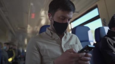 Siyah maskeli beyaz adam kulaklıkla müzik dinler ve trende seyahat ederken akıllı telefon kullanır. Toplu taşıma araçlarıyla sosyal mesafeler. Güvenli yaşam tarzı kavramı. COVID-19 salgını