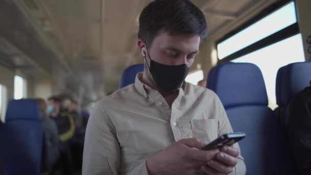 Hombre pasajero con máscara facial y auriculares usando el teléfono para ver videos en línea y leer noticias mientras viaja de seguridad en tren. Protección antivirus y uso de máscara en el transporte público. Nueva normalidad — Vídeos de Stock