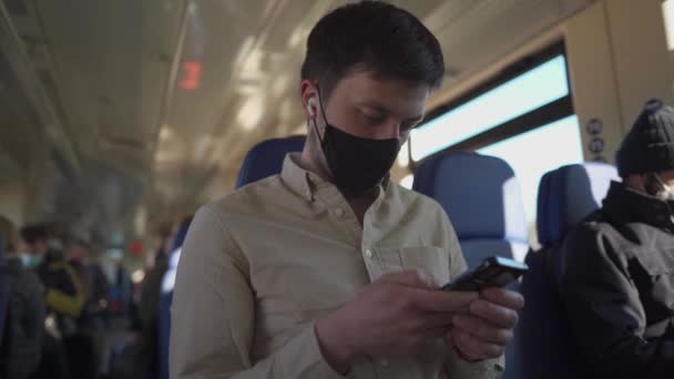 Veilig reizen. Man in beschermende masker en koptelefoon luisteren muziek op mobiele telefoon tijdens de trein reizen naar het werk tijdens coronavirus pandemie. Man gemaskerde gebruik oordopjes met smartphone in het openbaar vervoer — Stockvideo