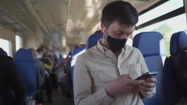 Hombre pasajero con máscara facial y auriculares usando el teléfono para ver videos en línea y leer noticias mientras viaja de seguridad en tren. Protección antivirus y uso de máscara en el transporte público. Nueva normalidad — Vídeos de Stock