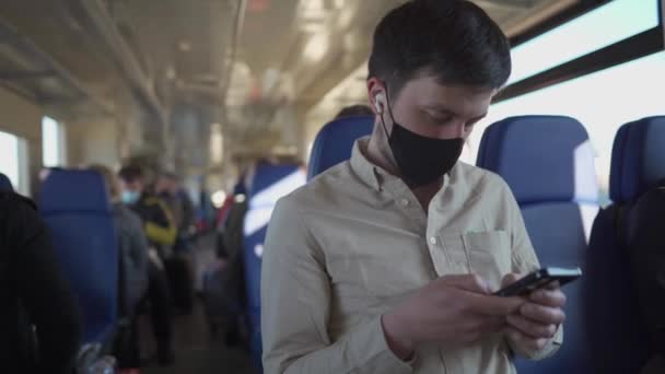 Hombre caucásico con máscara negra escucha música en los auriculares y utiliza el teléfono inteligente mientras viaja en tren. Distancia social viajando en transporte público. Concepto de estilo de vida de seguridad. pandemia de COVID-19 — Vídeos de Stock