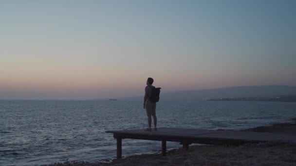 Silhouette di uomo viaggiatore con zaino godendo il tramonto sul molo di legno con vista sul mare mediterraneo sull'isola di Cipro, città di paphos. Concetto di viaggio. Backpacker sul molo di legno con vista sull'oceano — Video Stock