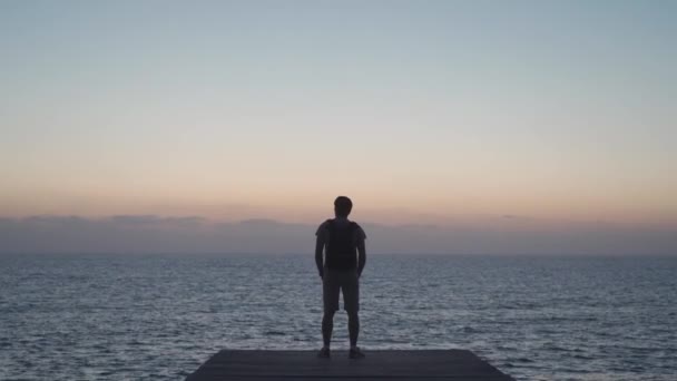 Silhouet van de mens reiziger met rugzak genieten van zonsondergang op houten pier met uitzicht op de Middellandse Zee op het eiland Cyprus, stad van paphos. Reisconcept. Backpacker op houten steiger met uitzicht op de oceaan — Stockvideo