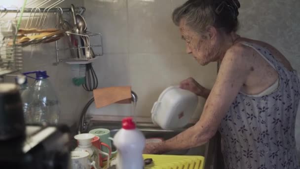 Pauvre femme âgée avec des émotions tristes, cheveux gris et rides profondes, 90 ans, lave et essuie la vaisselle dans la vieille cuisine. Triste femme âgée nettoie la vaisselle solitaire dans la petite cuisine. Pauvreté et misère — Video