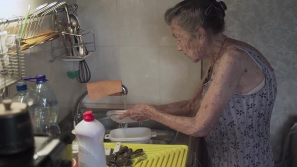 Eine ältere Frau spült Geschirr in der Küche. Traurige reife Hausfrau räumt in der Küche mit dem alten Geschirr auf. Empörte Frau mit grauen Haaren spült 90 Jahre alte Küchenutensilien in Handwäsche zu Hause — Stockvideo