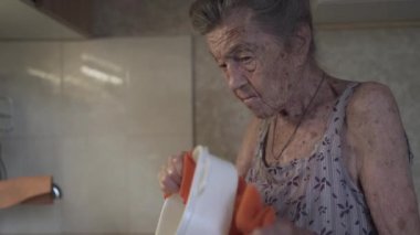 Mutfakta bulaşık yıkayan yaşlı bir kadın. Üzgün olgun ev hanımı mutfaktaki eski bulaşıkları yıkar. Gri saçlı üzgün bir kadın. 90 yaşında. Mutfak aletlerini evde yıkıyor.