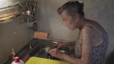 Zavallı yaşlı kadın, üzgün duygular, gri saçlar ve derin kırışıklıklar, 90 yaşında, eski mutfakta bulaşıkları yıkıyor ve siliyor. Üzgün yaşlı kadın küçük mutfakta tek başına çanak çömleği temizliyor. Yoksulluk ve sefalet