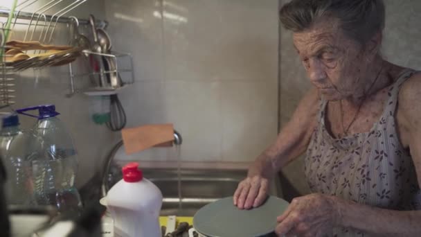 Mujer anciana solitaria y triste de 90 años de edad con el pelo gris obligada a lavar los platos con las manos debido a la pobreza en casa en una cocina vieja. Abuela en el trabajo. Antigua casa sucia, malas condiciones de vida — Vídeos de Stock