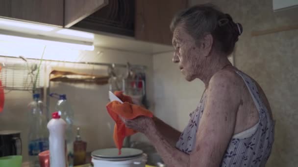 Eine einsame, traurige Seniorin von 90 Jahren mit grauen Haaren, die aufgrund der Armut zu Hause in einer alten Küche gezwungen ist, Geschirr mit den Händen zu waschen. Großmutter bei der Arbeit. Altes dreckiges Haus, schlechte Lebensbedingungen — Stockvideo