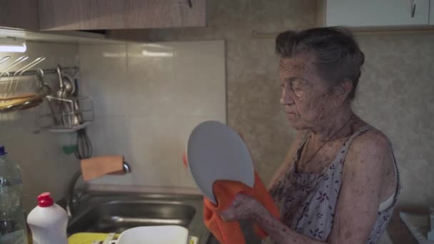 Szegény idős nő szomorú érzelmekkel, ősz hajjal és mély ráncokkal, 90 éves, mosogatja és törli az edényeket a régi konyhában. Szomorú idős nő takarít edények magányos kis konyhában. Szegénység és nyomorúság — Stock videók