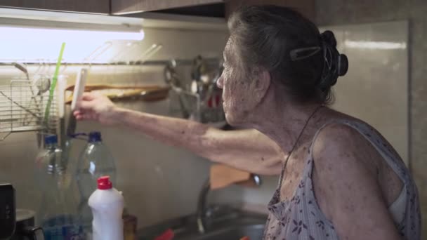 Lonely sad senior woman 90 years old with gray hair forced to wash dishes with her hands due to poverty at home in an old kitchen. Grandmother at work. Old dirty house, poor living conditions — Stock Video