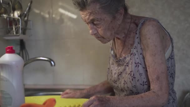 Eine ältere Frau spült Geschirr in der Küche. Traurige reife Hausfrau räumt in der Küche mit dem alten Geschirr auf. Empörte Frau mit grauen Haaren spült 90 Jahre alte Küchenutensilien in Handwäsche zu Hause — Stockvideo