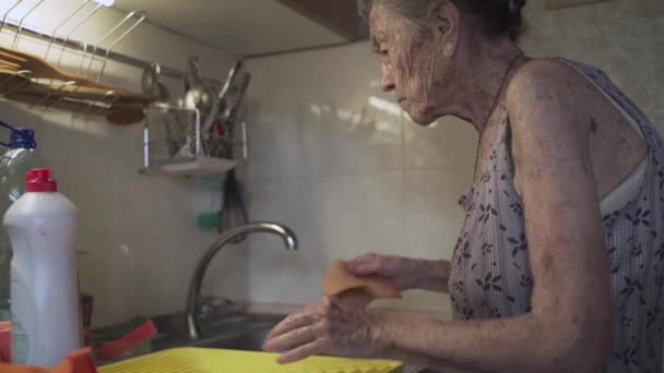 Samotna, smutna starsza kobieta, 90 lat, z siwymi włosami zmuszona zmywać naczynia rękami z powodu ubóstwa w domu, w starej kuchni. Babcia w pracy. Stary brudny dom, kiepskie warunki życia — Wideo stockowe