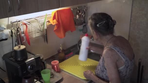 Pobre anciana con emociones tristes, canas y arrugas profundas, 90 años, lava y limpia platos en la cocina vieja. Triste anciana hembra limpia vajilla sola en la pequeña cocina. Pobreza y miseria — Vídeos de Stock