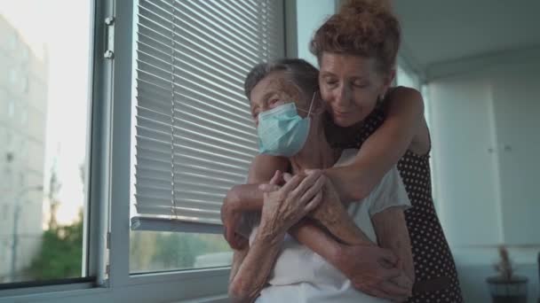 Femme âgée solitaire de 90 ans portant un masque médical et sa fille mature debout dans un étreinte regardant par la fenêtre dans une maison de retraite. Adulte fille visite mère âgée l'aime beaucoup et soutient — Video
