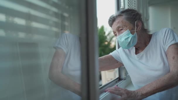 Retrato de una anciana enmascarada a finales de los 90 saludando desde la ventana del hogar. Mujer mayor con máscara protectora mira por la ventana y agita la mano mientras se aísla en casa y lleva a cabo distanciamiento social — Vídeos de Stock