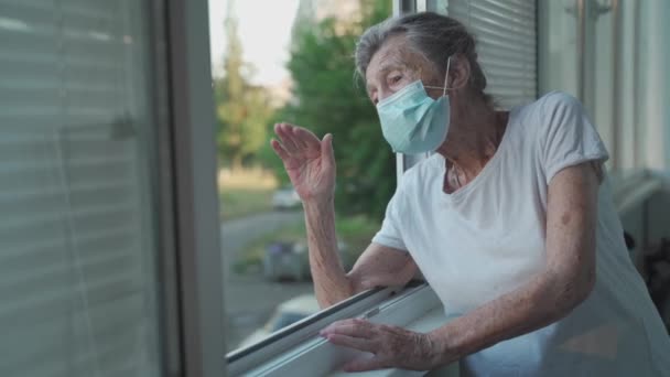 Una anciana solitaria enmascarada se para junto a la ventana y saluda con la mano a la familia que vino a visitarla al hospital. Mujer de 90 años disfrazada se despide de sus familiares en un asilo de ancianos — Vídeos de Stock