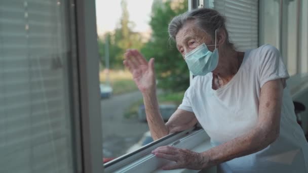 Porträtt av maskerad äldre kvinna i slutet av 90-talet vinkar från fönstret. Äldre kvinna i skyddsmask tittar ut genom fönstret och viftar med handen medan hon isolerar hemma och utför sociala distanser — Stockvideo