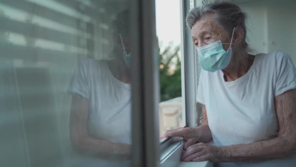 90 'ların sonlarında maskeli yaşlı bir kadının portresi ev penceresinden el sallıyor. Koruyucu maskeli yaşlı kadın pencereden dışarı bakar ve evde yalnızken elini sallar ve sosyal mesafeyi yönetir. — Stok video