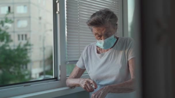 Femme âgée appliquant un désinfectant pour les mains, portant un masque de protection contre Covid-19 debout près de la fenêtre. Femme âgée triste solitaire dans le masque médical du visage à l'aide d'un désinfectant liquide antibactérien à la maison — Video