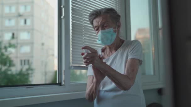 Üst düzey kadın el dezenfektanı sürüyor, Covid-19 'a karşı koruyucu maske takıyor, pencerenin önünde duruyor. Evde el antibakteriyel sıvı dezenfektanı kullanan, yüzü maskeli, üzgün, yaşlı bir kadın. — Stok video