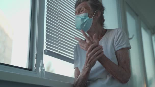 Старшая женщина применяет дезинфицирующее средство для рук, надевает защитную маску против Ковид-19, стоящего у окна. Одинокая грустная пожилая женщина в маске для лица с помощью антибактериального средства для дезинфекции рук дома — стоковое видео
