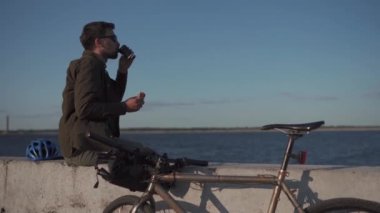 Bisikletçi deniz kenarındaki beton çitte oturur ve tek kullanımlık kahveden içer ve bisiklet gezisinde sandviç yer. Adam göl kenarında bisiklet sürdükten sonra öğle yemeği yiyor. İşten sonra büyük bir gölete bakan atıştırmalıklar.