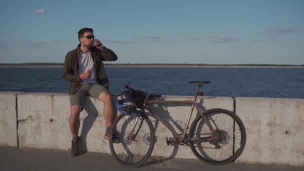 Erkek bisikletçi, deniz kenarında gezinirken sandviç yer ve kahve içer. İnsan büyük göl kenarında bisiklet sürdükten sonra abur cubur yer ve sıcak içecek içer. Kahveli yemek. Yemek molası. — Stok video