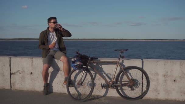 Homme cycliste mange sandwich et boit du café pour aller tout en étant assis sur la promenade par la mer. L'homme mange des collations et boit des boissons chaudes après avoir fait du vélo près du grand lac. Plats à emporter avec du café. Pause déjeuner — Video