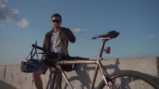 Ciclista senta-se em cerca de concreto perto do mar e bebidas de xícara descartável de café e come sanduíche em viagem de bicicleta. O homem almoça depois de andar de bicicleta perto do lago. Lanche após o trabalho com vista para uma grande lagoa — Vídeo de Stock