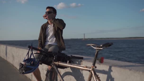 Un uomo con la bicicletta che fa colazione sul lungomare. Pranzo all'aperto dopo il ciclismo sul terrapieno dal mare. Rifornimento con panino e caffè prima di andare al lavoro a ruota con vista sul grande lago — Video Stock