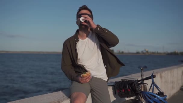 Homem com bicicleta tomando café da manhã na beira-mar. Almoço ao ar livre depois de andar de bicicleta em aterro por mar. Reabastecendo com sanduíche e café antes do passeio para trabalhar uma roda com vista para o grande lago — Vídeo de Stock