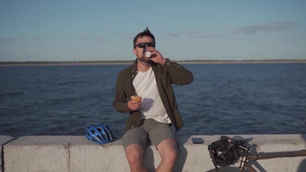 男性サイクリストは海沿いの遊歩道に座っている間に行くためにサンドイッチやドリンクコーヒーを食べます。男は大きな湖でサイクリングした後に軽食を食べて熱い飲み物を飲む。コーヒーとテイクアウト食品。昼休み — ストック動画
