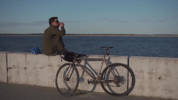 Mand cyklist spiser sandwich og drikker kaffe til at gå, mens du sidder på promenaden ved havet. Mand spiser snack og drikker varm drikke efter cykling ved stor sø. Takeaway mad med kaffe. Frokostpause – Stock-video