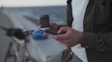 Bir adamın el yapımı kahve içerken ve deniz kenarında bisikletle dolaşırken cep telefonu kullanırken yakın çekimi. Eko taşıma ve kahve molası. Bisiklet teslimatı ara verdi. 4k video