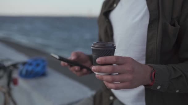 Leyendo noticias, redes sociales, navegando por Internet. 5G en su teléfono inteligente. Descanso. Primer plano del ciclista masculino de mano sosteniendo el teléfono inteligente mientras bebe café para llevar cerca de la bicicleta en el paseo marítimo — Vídeos de Stock