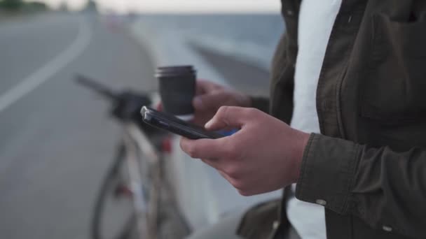 Unrecognizable ciclista maschile utilizzare smartphone e bevande da asporto caffè mentre in piedi vicino alla bicicletta su argine. Consegna uomo in bicicletta durante la pausa caffè sulla pista ciclabile via mare. Social network outdoor — Video Stock