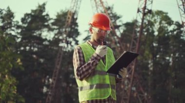 Elektrik mühendisi, başlık ve güvenlik yeleği giyiyor, yüksek voltajlı elektrik hatlarında elektrik sistemlerini kontrol ediyor, telsizle konuşuyor, müfettişin çalışma planı panosu var. Elektrik şirketi çalışanı