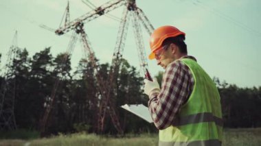 Koruyucu üniformalı ve turuncu şapkalı beyaz mühendis, arka planda elektrik iletim hattının telsiz ve panosunu tutuyor. Yüksek voltaj kulesinin yanında telsizi olan elektrik işçisi.