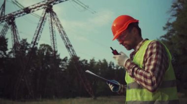 Koruyucu üniformalı ve turuncu şapkalı beyaz mühendis, arka planda elektrik iletim hattının telsiz ve panosunu tutuyor. Yüksek voltaj kulesinin yanında telsizi olan elektrik işçisi.