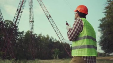 Elektrik mühendisi, başlık ve güvenlik yeleği giyiyor, yüksek voltajlı elektrik hatlarında elektrik sistemlerini kontrol ediyor, telsizle konuşuyor, müfettişin çalışma planı panosu var. Elektrik şirketi çalışanı