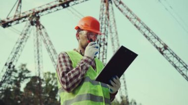 Koruyucu üniformalı ve turuncu şapkalı beyaz mühendis, arka planda elektrik iletim hattının telsiz ve panosunu tutuyor. Yüksek voltaj kulesinin yanında telsizi olan elektrik işçisi.