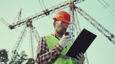 Elektrik mühendisi, başlık ve güvenlik yeleği giyiyor, yüksek voltajlı elektrik hatlarında elektrik sistemlerini kontrol ediyor, telsizle konuşuyor, müfettişin çalışma planı panosu var. Elektrik şirketi çalışanı