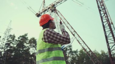 Koruyucu üniformalı ve turuncu şapkalı beyaz mühendis, arka planda elektrik iletim hattının telsiz ve panosunu tutuyor. Yüksek voltaj kulesinin yanında telsizi olan elektrik işçisi.
