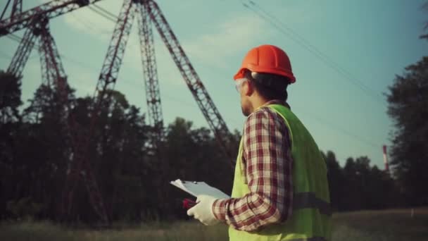 Electrical equipment worker near high voltage tower using walkie talkie and clipboard. Industrial engineer and power lines and towers. Supervising electrification near transmission line outdoors — Stock Video